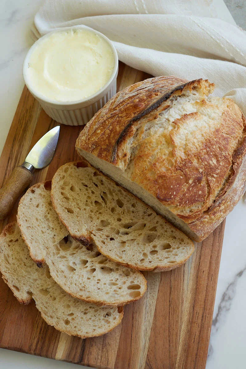 A golden sourdough loaf sitting on a wooden cutting board with a linen towel beside it