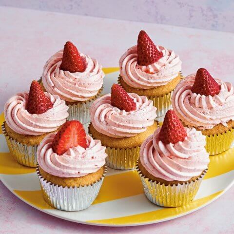 Seven vanilla cupcakes with pink swirled frosting and a strawberry on top arranged on a plate