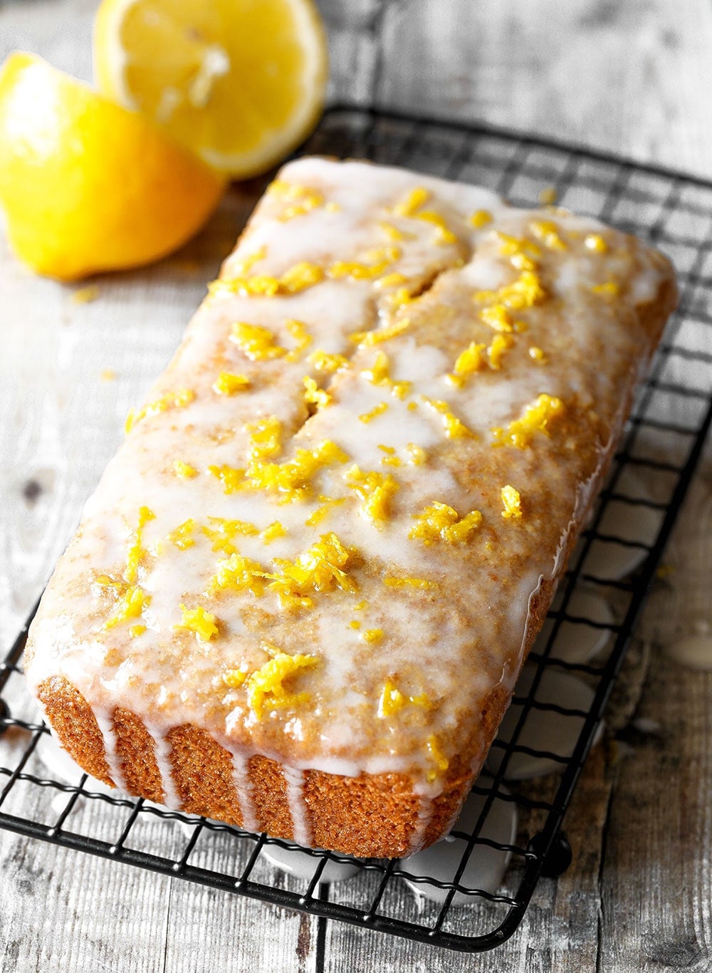 A lemon drizzle cake on a wire cooling rack with white glaze dripping down the sides and lemon slices beside it