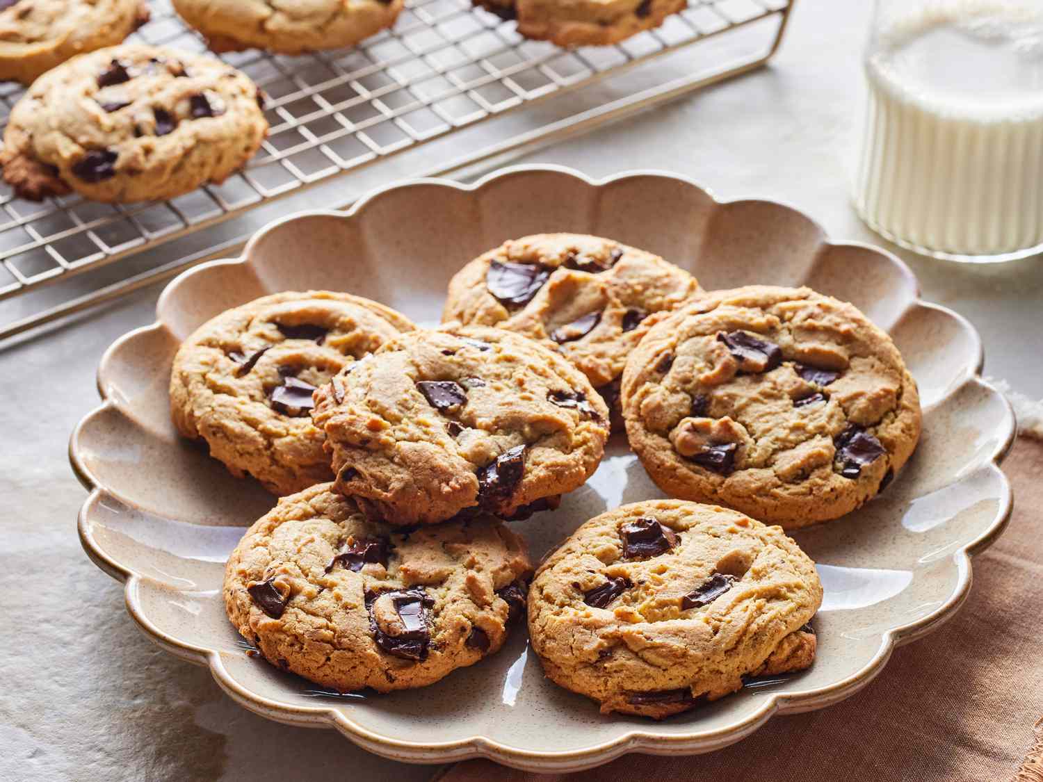 Six chocolate chip cookies arranged on a white plate with a glass of milk in the background