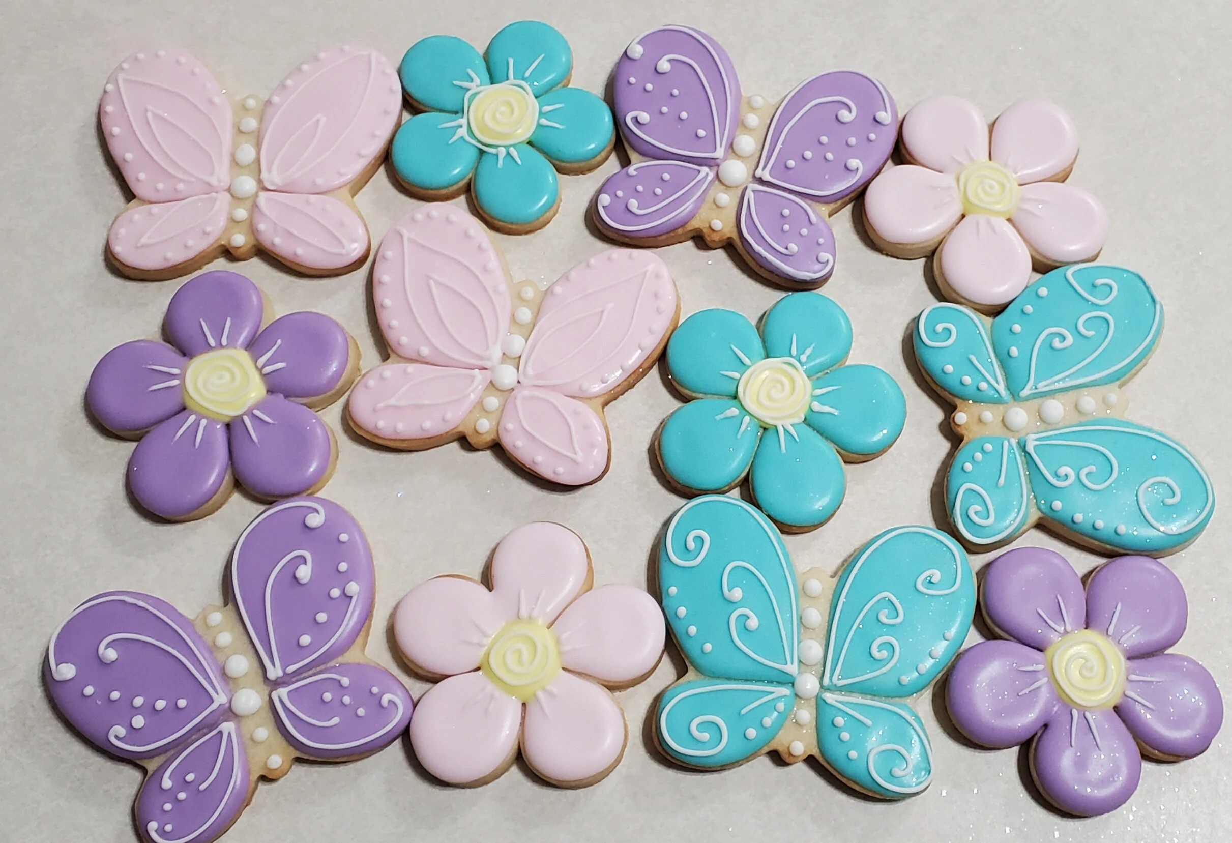 A dozen decorated sugar cookies in pastel colors shaped like flowers and butterflies on a white tray